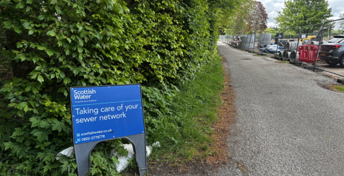 A Scottish Water sign reads 'Taking care of your sewer network', with green bushes behind it and a tarmac single lane road to the right