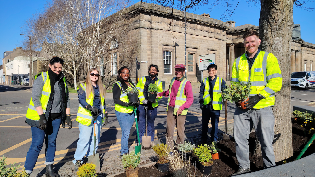A group of seven people wearing high-visibility vests and gloves are planting greenery around a tree in an urban setting. They are standing on a paved area with a mix of potted plants, soil, and gardening tools. The background features a historic stone building with large windows and columns, as well as a clear blue sky. The group appears cheerful and engaged in their community gardening activity.