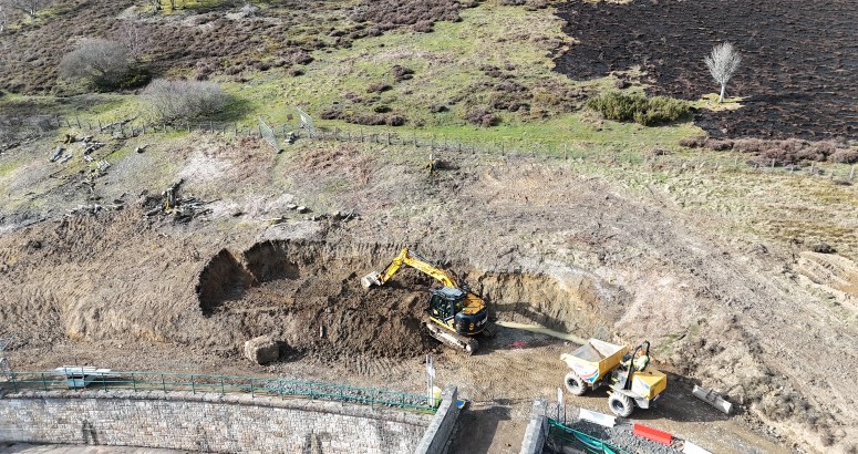 Aerial view of construction site with an excavator and a loader working on a large dirt area. The surrounding landscape includes grassy and rocky patches.