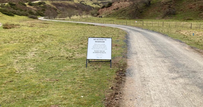 A gravel path curves through a grassy landscape with gently rolling hills in the background. A sign by the side of the path reads, "No access to reservoir due to ongoing works."