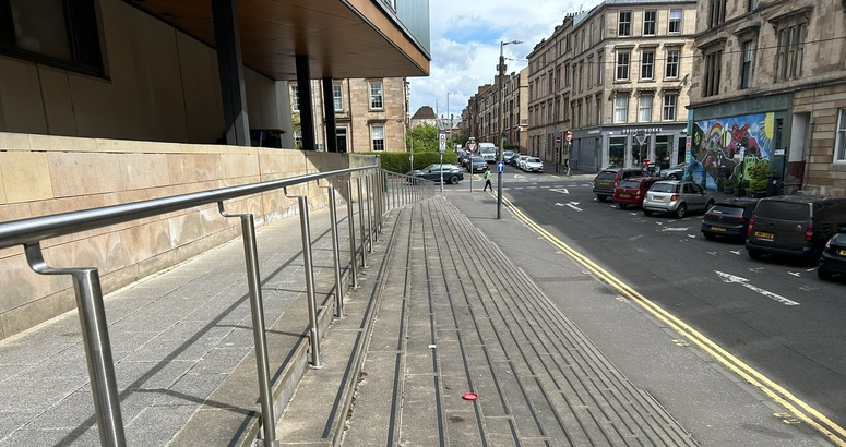 A gently sloping concrete access ramp with stainless steel handrails runs alongside a modern building on an urban street. Stone tenement buildings, parked cars, and a colourful mural are visible across the road under a partly cloudy sky.