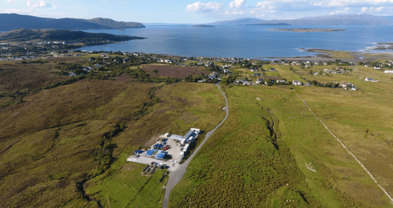 A drone shot of green landscape with a works site, with the coastline in the background.