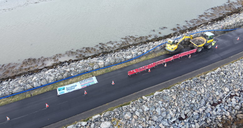 Aerial view of road construction near a water body with vehicles and equipment, featuring a "Caledonia Water Alliance" banner.