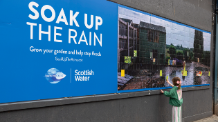 Large billboard on a city street promoting Scottish Water’s “Soak Up the Rain” campaign, featuring a photo of rainwater flooding a garden while a child reaches toward the display.