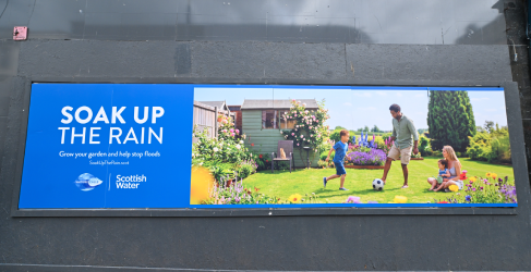 Wide billboard advertisement for Scottish Water’s “Soak Up the Rain” campaign, showing a bright garden area with grass and flowers and a family playing with a ball. 
