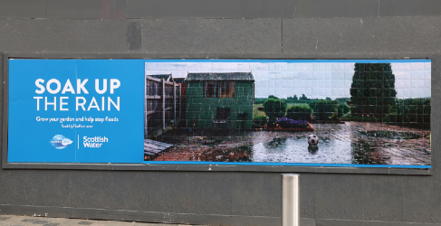 Wide billboard advertisement for Scottish Water’s “Soak Up the Rain” campaign, showing a flooded garden with the message encouraging people to grow gardens and help stop floods.