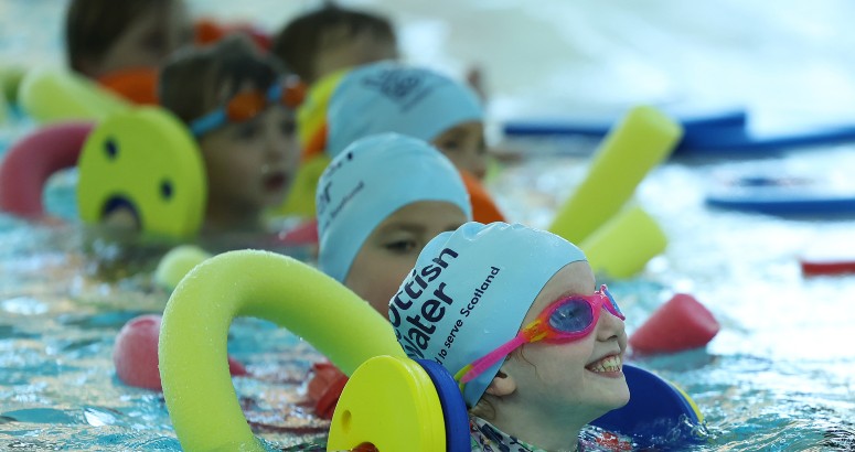 Children wearing Scottish Water Blue swimming caps use colourful pool noodles during a swimming lesson
