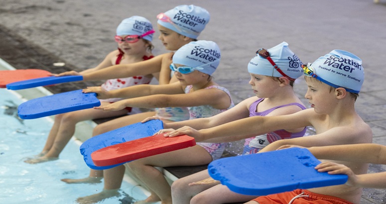 5 kids sitting at edge of pool with blue swim caps on and holding out a mix of red and blue floats.