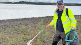 Ed the Ranger with his litter picker and rubbish bag collecting litter with reservoir in background. 