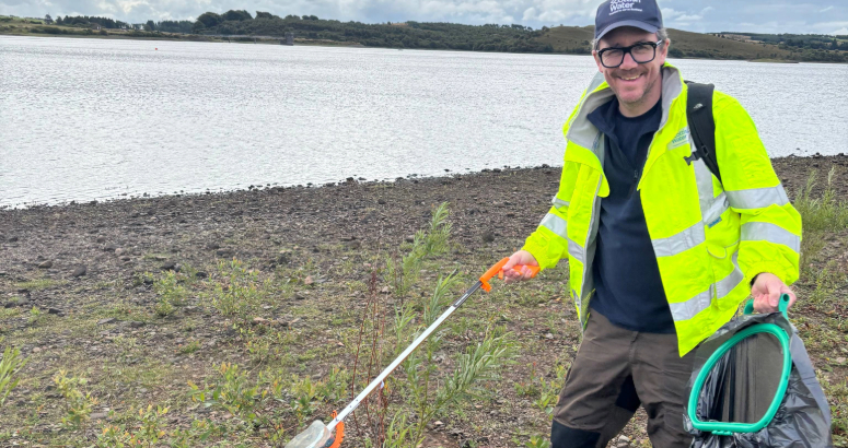 Ed the Ranger with his litter picker and rubbish bag collecting litter with reservoir in background. 