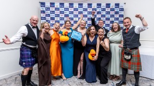 A group of 10 members of West Dunbartonshire Leisure at the awards night, with 3 men dressed in kilts and 7 ladies in dresses. They are cheering with women in middle of group holding the award.
