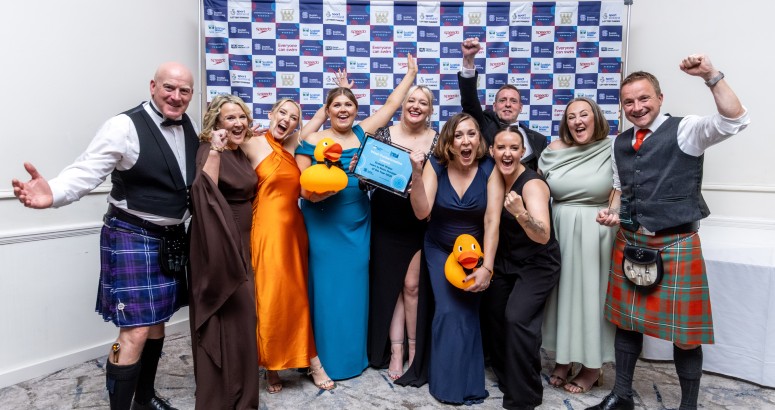 A group of 10 members of West Dunbartonshire Leisure at the awards night, with 3 men dressed in kilts and 7 ladies in dresses. They are cheering with women in middle of group holding the award.