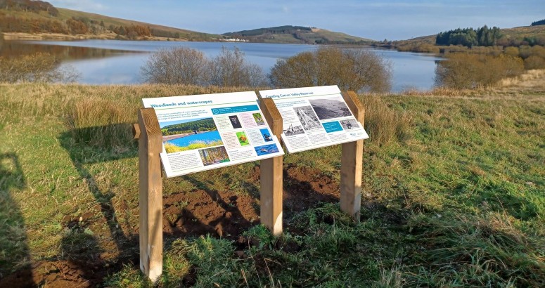 2 new signs side by side at Carron Valley on a sunny day with reservoir in background.
