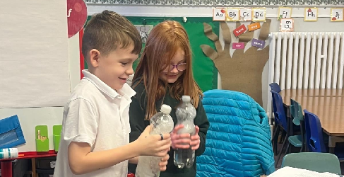 Two children in a classroom hold plastic bottles and smile as they engage in an activity. The background features colorful educational decorations, chairs, and tables.