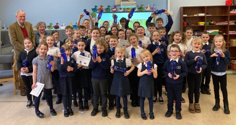 Group of schoolchildren proudly displaying Scottish Water reusable water bottles in a classroom, with an adult supervising in the background.