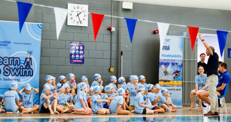Group of children in swim caps seated on the poolside during a swimming class at a public aquatic facility, with instructors standing by and promotional banners visible in the background.